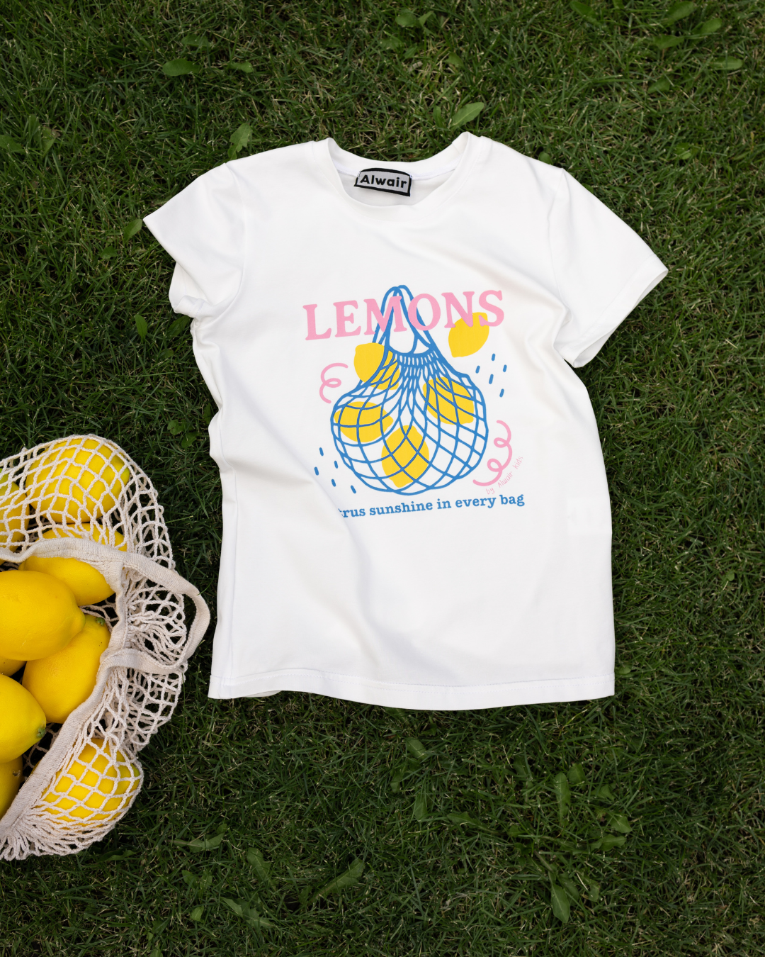  white kids t-shirt with lemon print lying on grass next to yellow lemons Alwair Kids summer collection