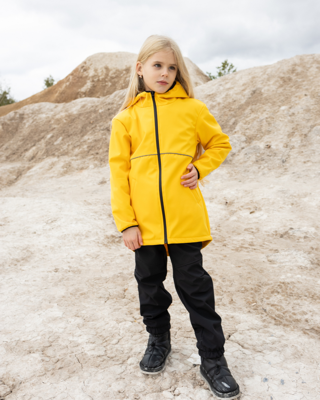 Girl wearing yellow softshell jacket for kids by Alwair Kids on rocky background

