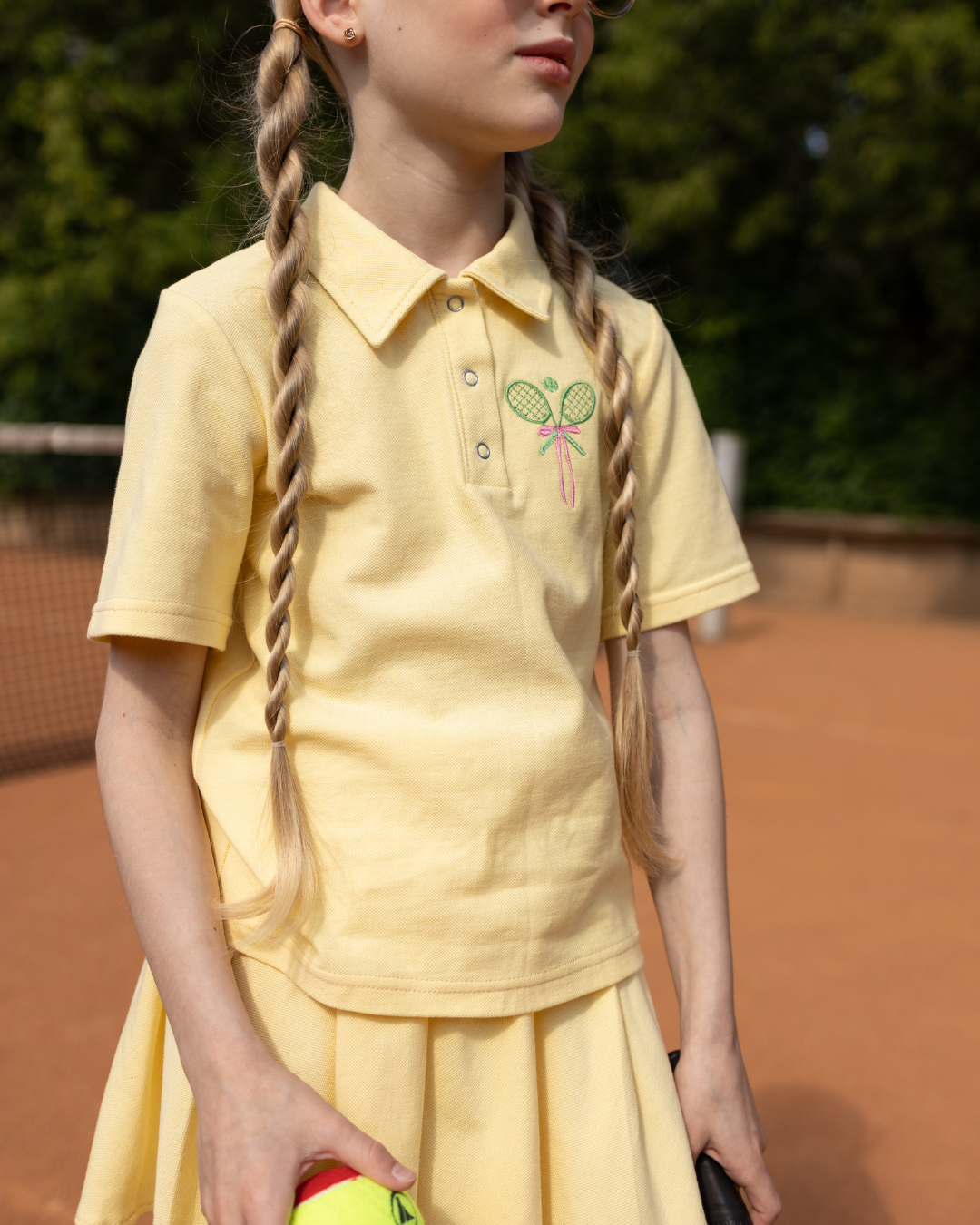 Girl wearing yellow tennis polo shirt by Alwair Kids with embroidered rackets on a tennis court