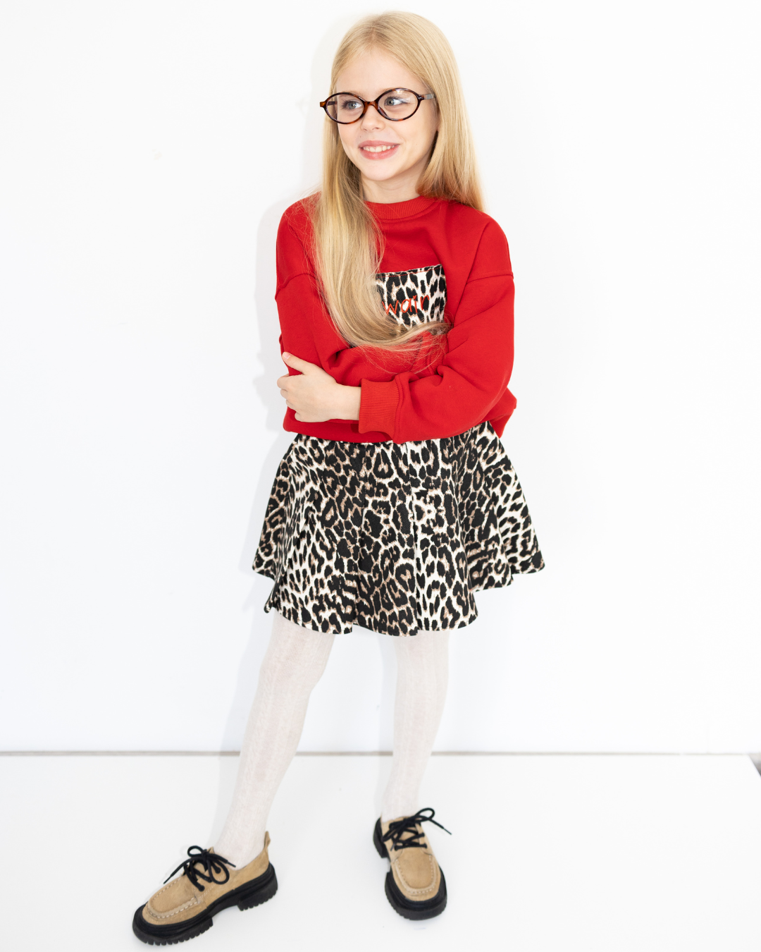 Smiling girl in leopard print skirt and red sweatshirt from Leo capsule by Alwair Kids, standing with arms crossed
