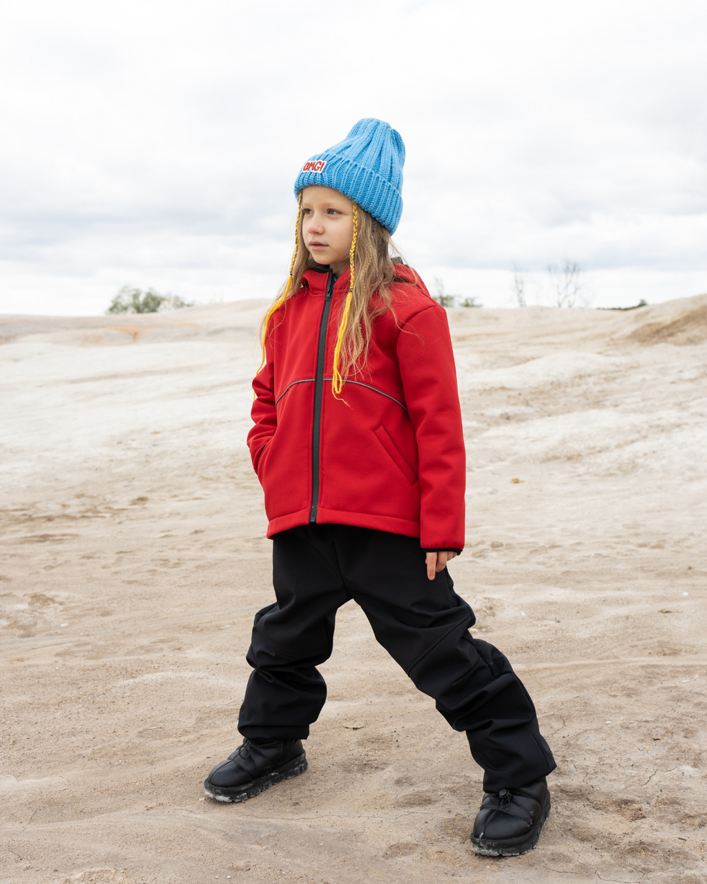 Kid in red softshell jacket and blue hat by Alwair Kids standing on sandy terrain

