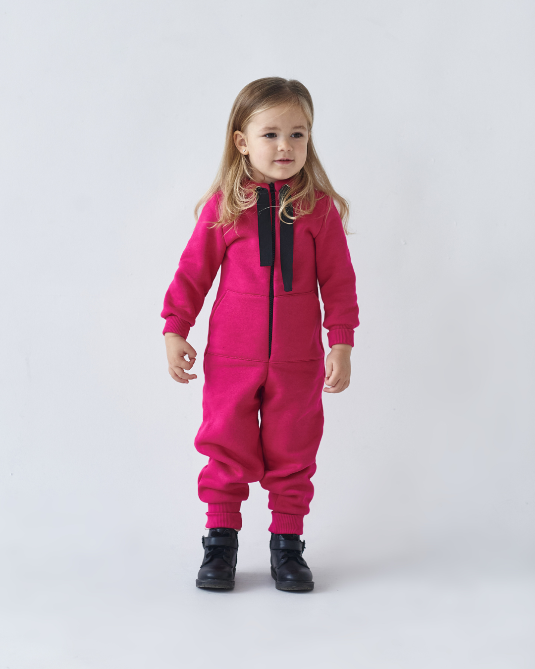 Toddler girl wearing raspberry pink fleece jumpsuit by Alwair Kids, standing in studio with black boots
