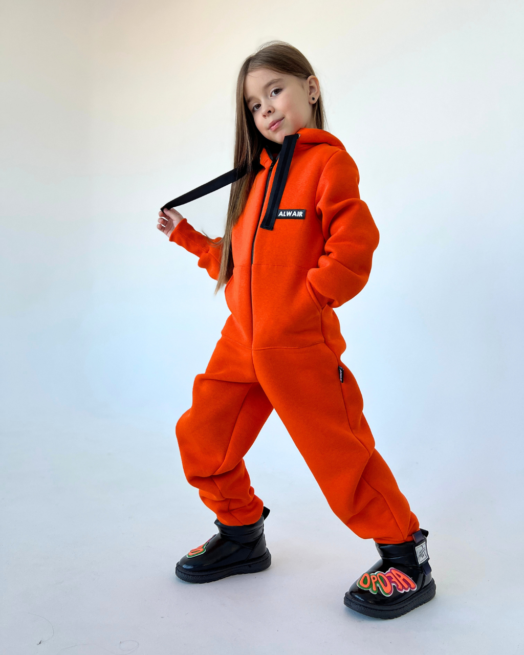 Girl wearing bright orange fleece jumpsuit by Alwair Kids with hood and black boots, posing confidently in studio