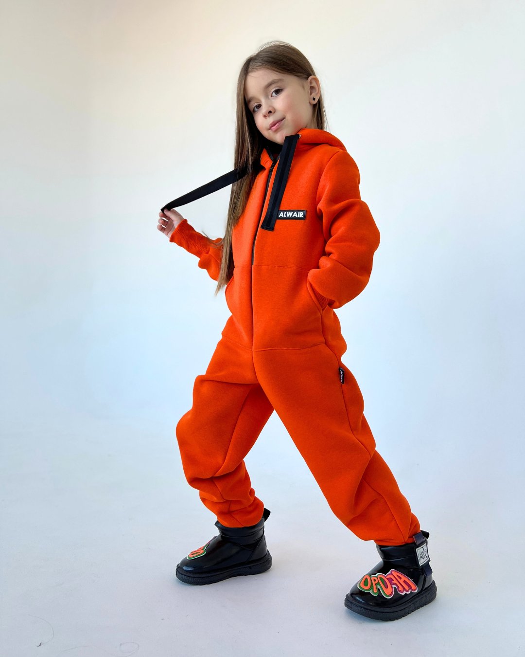 Girl wearing bright orange fleece jumpsuit by Alwair Kids with hood and black boots, posing confidently in studio