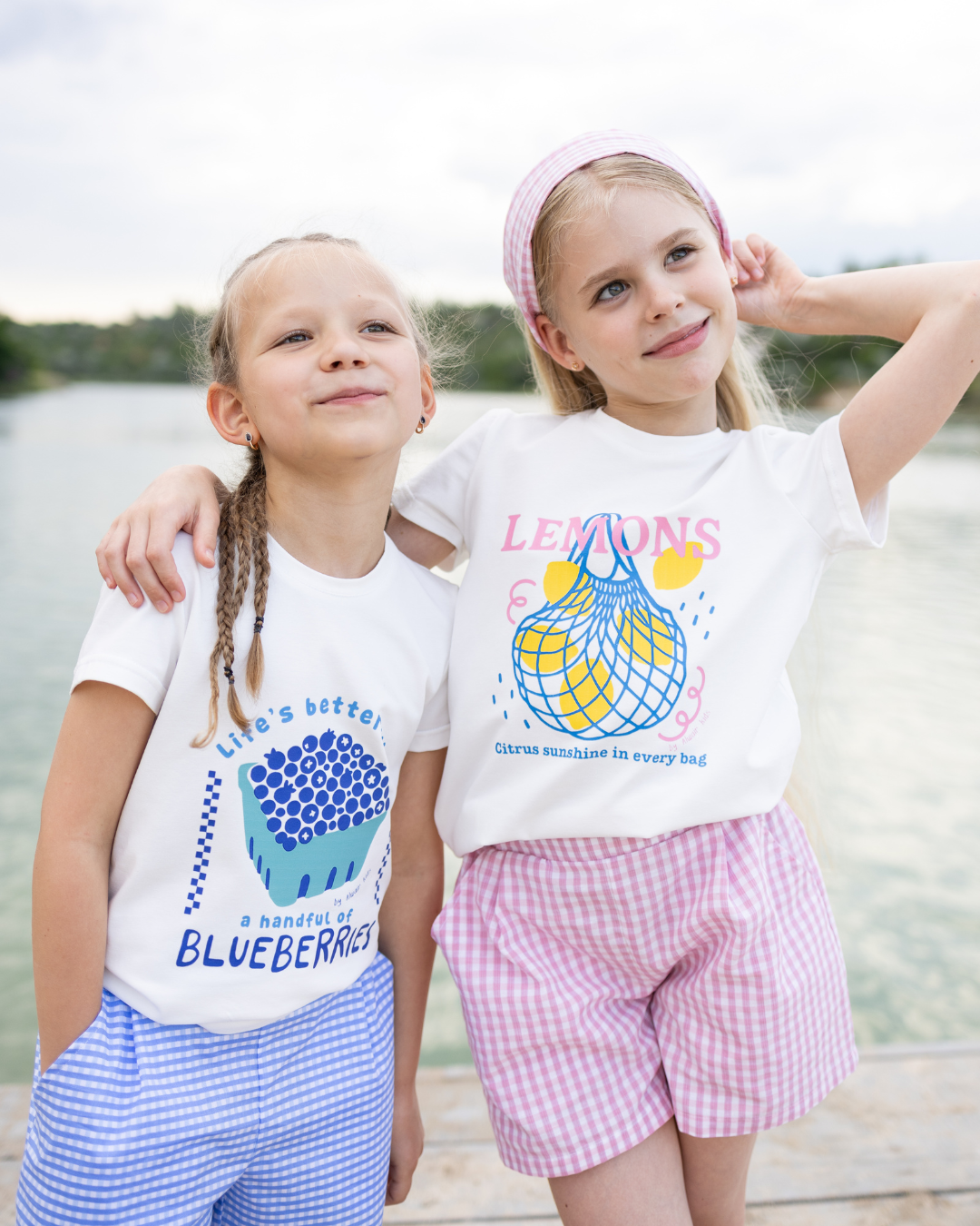Two girls wearing summer print t-shirts – one with 'Blueberries' and blue gingham shorts, the other with 'Lemons' and pink gingham shorts – cheerful kidswear look by the lake