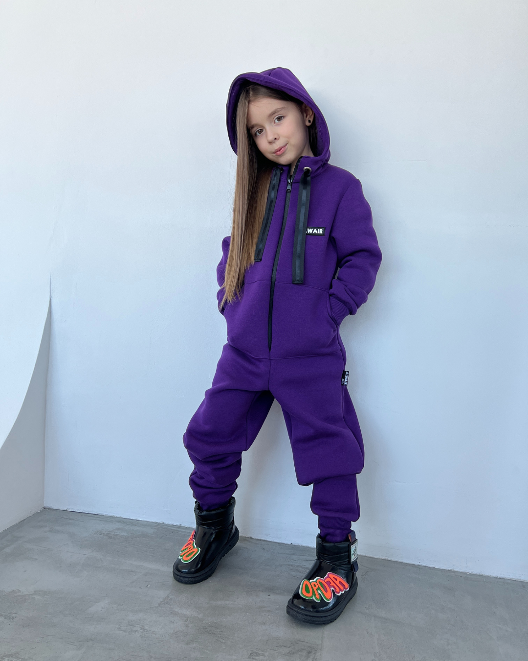 Girl in purple fleece jumpsuit by Alwair Kids with hood and black boots, standing against white wall